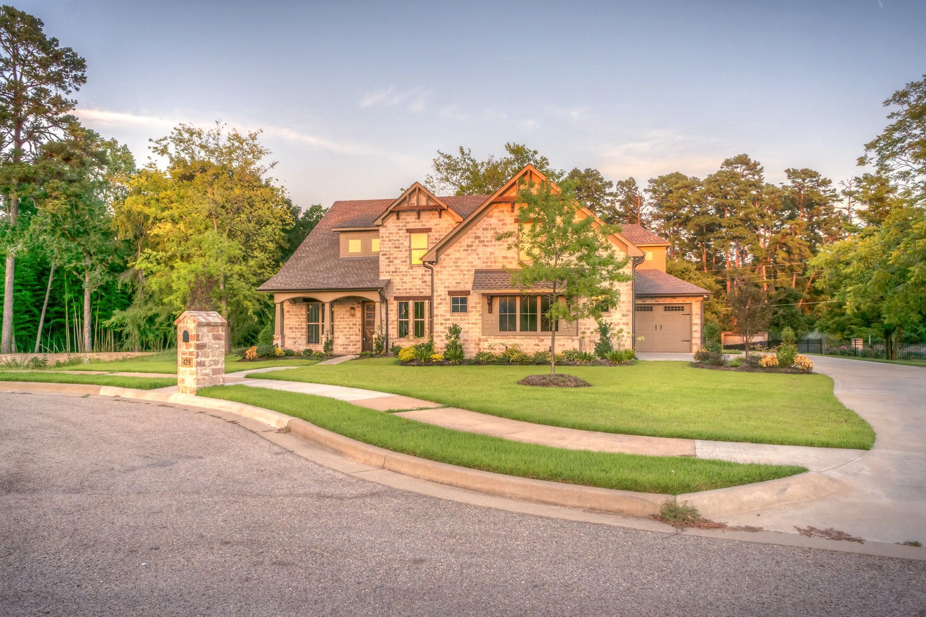 Elegant stone family home with manicured lawn, trees, and driveway on a sunny day.